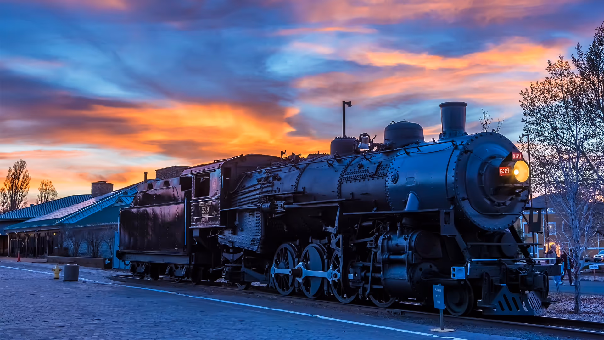 Black steam locomotive train parked on railway track at sunset with vibrant orange and blue sky.
